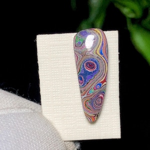 May include: A polished, teardrop-shaped gemstone with a swirling pattern of colours including purple, blue, yellow, and red. The stone is mounted on a white surface, showcasing its unique, marbled appearance. The gemstone is approximately 5 cm long.