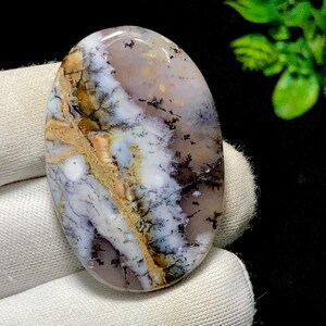 May include: An oval gemstone with a dendritic pattern. The stone displays a mix of colours, including white, brown, and black, with a translucent quality. The gemstone is polished and smooth, showcasing its natural beauty. Suitable for jewellery making.