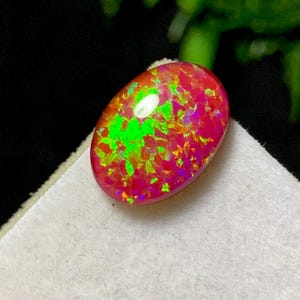 May include: Oval-shaped opal gemstone with a vibrant display of color. The stone features a base of deep red and pink hues, interspersed with flashes of bright green and orange. The opal is set against a neutral background.