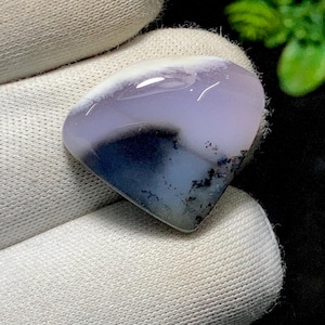 May include: A polished gemstone with an organic shape. The stone displays a mix of white, lavender, and navy blue hues, with detailed patterns and dark speckles. The gemstone is held in white fabric.