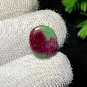 Könnte beinhalten: Ovaler Edelstein mit polierter Oberfläche. Der Stein zeigt eine lebendige Farbkombination aus tiefem Rot und hellem Grün. Der Edelstein wird vor einem weißen Stoffhintergrund präsentiert, der seine natürlichen Farben und Texturen hervorhebt.