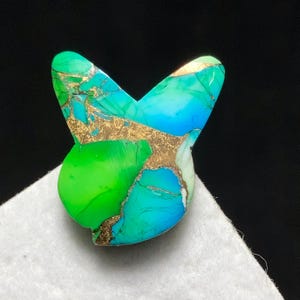 May include: A butterfly-shaped gemstone in shades of green, turquoise, and blue, with gold-coloured veining. The stone is set against a white surface and a black background. The gemstone has a unique, natural appearance.