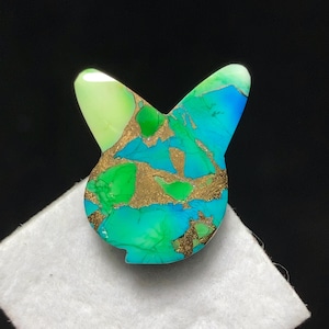 Puede incluir: Una gema colorida en forma de conejo, con una mezcla de tonos verdes, azules y dorados. La piedra está pulida y tiene una superficie lisa. La forma de conejo es un diseño único, perfecto para joyería o fines decorativos.