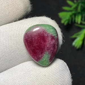 May include: A polished gemstone, teardrop-shaped, with a deep red centre and green edges. The gem is held in white fabric. The stone's colours are vivid and natural.