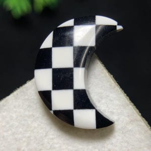 Puede incluir: Un objeto decorativo en forma de media luna con un patrón de cuadros blancos y negros. El objeto está hecho de un material brillante y tiene una superficie lisa. El diseño recuerda a un tablero de ajedrez. Es probable que sea un accesorio decorativo.