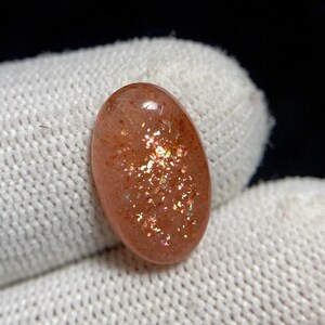 May include: Oval-shaped sunstone gemstone with a polished surface. The stone is a warm peach color with sparkling inclusions that catch the light. The gemstone is set against a white fabric background.