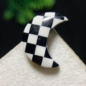 May include: A crescent-shaped accessory with a black and white checkered pattern. The object is set against a neutral background, highlighting its contrasting colors and unique design. The accessory's shape and pattern suggest a decorative or jewelry item.