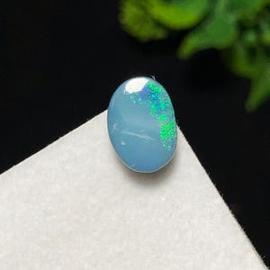 May include: Oval-shaped opal gemstone with a light blue base color. The opal has vibrant flashes of green and blue, creating a play-of-color effect. The gemstone is set against a white surface with a dark background.