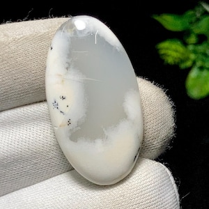 May include: An oval gemstone featuring a white and grey cloudy pattern with tiny black specks. The polished stone is displayed against a white fabric, highlighting its natural appearance. Suitable for jewellery making, this stone is a unique find.