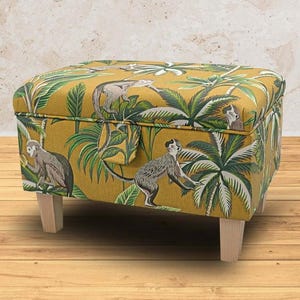 May include: Mustard yellow square ottoman with a jungle-themed fabric featuring monkeys and tropical plants. The ottoman has four wooden legs and a hinged lid. The fabric design incorporates green, grey, and white elements. The ottoman measures approximately 60cm x 60cm.