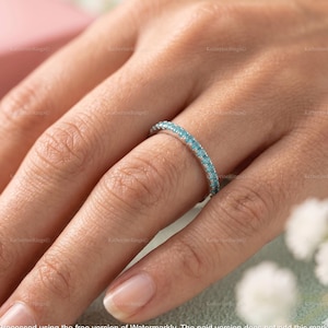 Könnte beinhalten: Ein silberner Ring mit einer Reihe kleiner, hellblauer Edelsteine. Der Ring befindet sich an einem Finger, wobei die Hand im Vordergrund ist. Der Hintergrund ist unscharf, mit einem Hauch von rosa Objekt und weißen Blumen.