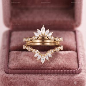 May include: A gold ring guard set featuring two bands with marquise and round cut white stones. The top band has a pointed arch design with marquise stones, while the bottom band mirrors this shape. Both bands have intricate metal detailing.