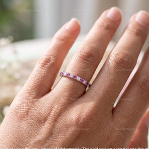 May include: A silver-toned band ring featuring an alternating pattern of round-cut pink gemstones and clear white stones set in a continuous row along the top of the band. The ring is worn on a finger, showcasing the circular arrangement of stones.