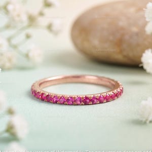 May include: A rose gold ring, its band set with bright pink gemstones. The ring sits on a pale green surface, with white flowers and a smooth, light brown stone in the background. A delicate piece of jewellery.