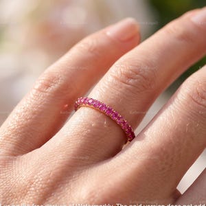 May include: A thin gold band ring featuring a continuous row of small, round, vibrant pink gemstones set in a shared prong setting. The ring encircles the finger, showcasing a uniform design with polished metal edges and bright, saturated stones.