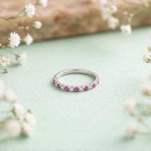 Könnte beinhalten: Ein dünner Silberring mit einem abwechselnden Muster aus runden rosa Edelsteinen und klaren weißen Diamanten, die entlang des gesamten Umfangs des Metallbandes gefasst sind und ein durchgehendes kreisförmiges Design aus funkelnden Steinen bilden.