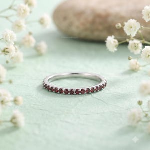 May include: A thin silver band ring featuring a continuous row of small, round, deep red garnet gemstones set in a shared prong setting. The ring has a polished metallic finish and a circular shape, showcasing a uniform design around the entire band.