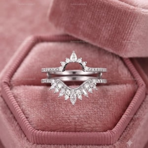 May include: A silver-toned ring set featuring two stacked bands. The top band has a curved arch with small round stones, while the bottom band displays a fan-like arrangement of marquise and round stones. Both bands are set with clear, faceted gems.