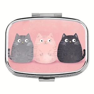 May include: A silver pill box with a pink background featuring three cartoon cats in gray, pink, and black. The cats have large eyes and are sitting side by side. The pill box has a rectangular shape with rounded edges.