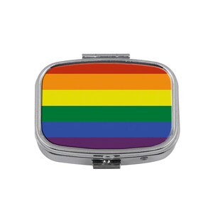 May include: A small, rectangular pill box with a silver-colored metal frame. The top of the box features the rainbow flag design, with horizontal stripes of red, orange, yellow, green, blue, and purple. The box is closed.