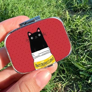 Puede incluir: Un pastillero rectangular con borde plateado y fondo rojo con puntos blancos. Presenta una ilustración de un gato negro saliendo de un frasco amarillo con la palabra "Antidepresivo".