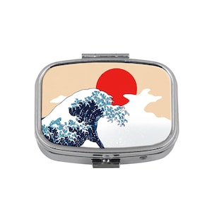 May include: A silver pill box with a rectangular shape. The lid features a Japanese-style design of a large ocean wave in shades of blue and white, a red sun, and a beige sky.