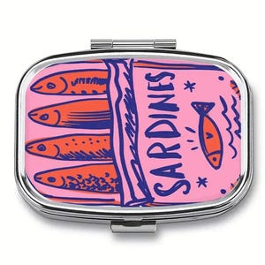 Pode incluir: Um estojo de comprimidos prateado com uma tampa rosa com um design de lata de sardinhas. O design inclui sardinhas laranja, contornos azuis e a palavra "SARDINES" em azul. Uma pequena ilustração de peixe laranja também está presente.