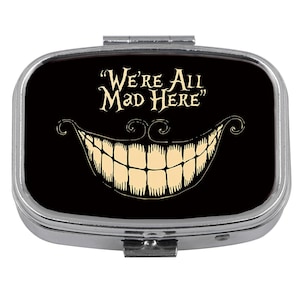 Könnte beinhalten: Eine silberne Pillendose mit schwarzem Hintergrund, die ein grinsendes Cheshire-Cat-Lächeln und den Text "We're All Mad Here" in einer skurrilen Schriftart zeigt. Die Dose hat eine rechteckige Form.