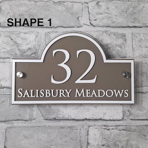 May include: A rectangular house number sign with a brown background and white trim. The number "32" is prominently displayed in white, along with the text "SALISBURY MEADOWS". The sign is mounted on a brick wall. The text "SHAPE 1" is at the top.