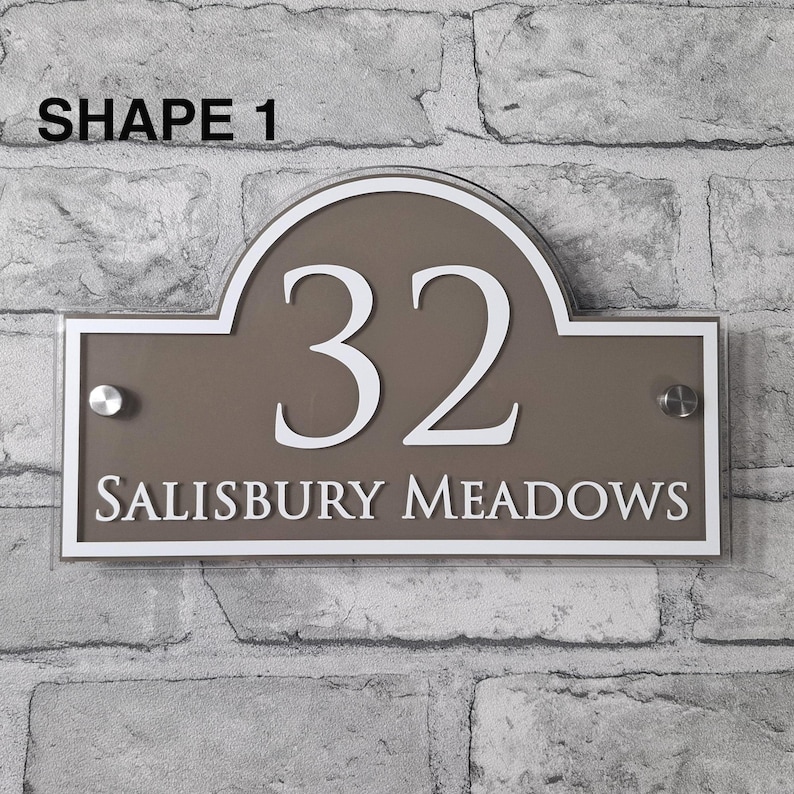 May include: A rectangular house number sign with a brown background and white trim. The number "32" is prominently displayed in white, along with the text "SALISBURY MEADOWS". The sign is mounted on a brick wall. The text "SHAPE 1" is at the top.