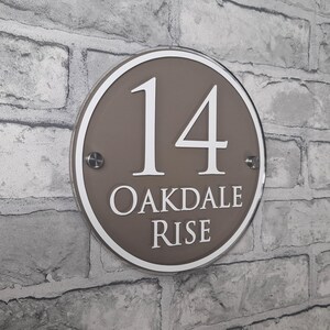 May include: A round, brown house number sign with the number 14 in white, along with the words "OAKDALE RISE". The sign is mounted on a gray brick wall. The sign has a white border and is secured with two silver screws.