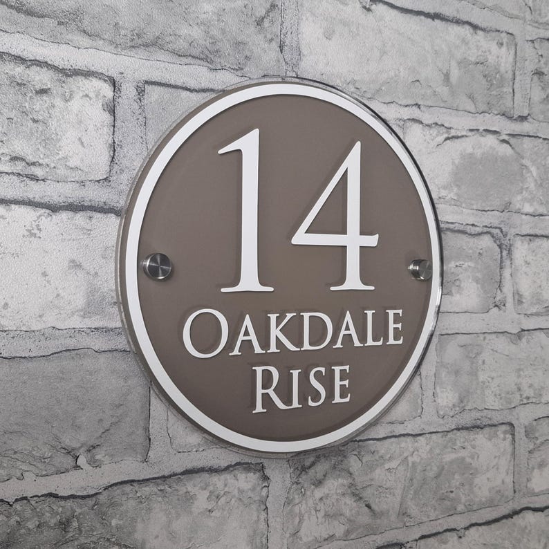 May include: A round, brown house number sign with the number 14 in white, along with the words "OAKDALE RISE". The sign is mounted on a gray brick wall. The sign has a white border and is secured with two silver screws.