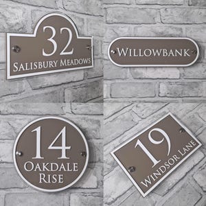 May include: Four house number signs in various shapes. Each sign is taupe with white text. The signs read "32 SALISBURY MEADOWS", "WILLOWBANK", "14 OAKDALE RISE", and "19 WINDSOR LANE". The signs are mounted on a gray brick wall.