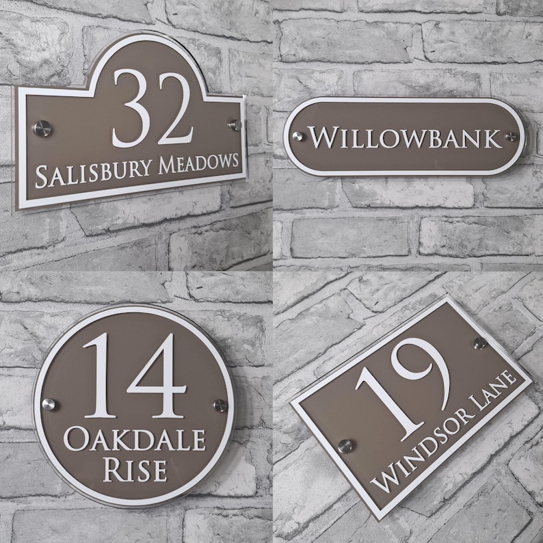 May include: Four house number signs in various shapes. Each sign is taupe with white text. The signs read "32 SALISBURY MEADOWS", "WILLOWBANK", "14 OAKDALE RISE", and "19 WINDSOR LANE". The signs are mounted on a gray brick wall.