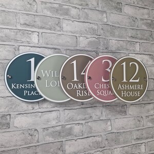 May include: Five round address signs in various colors, including teal, sage green, brown, rose, and olive green. Each sign displays a number and street name in white lettering. The signs are mounted on a brick wall.