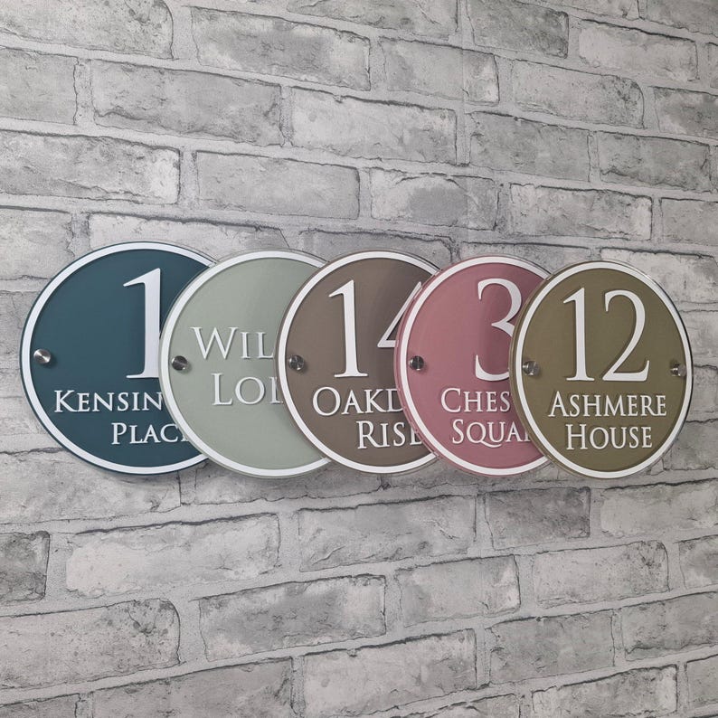 May include: Five round address signs in various colors, including teal, sage green, brown, rose, and olive green. Each sign displays a number and street name in white lettering. The signs are mounted on a brick wall.