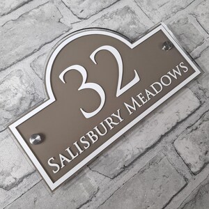 May include: A rectangular house number sign with a taupe background and white trim. The number "32" is prominently displayed in white, above the words "SALISBURY MEADOWS". The sign is mounted with two silver screws.