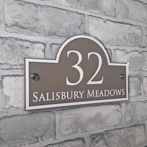 May include: A rectangular house number sign with the number "32" in white, above the words "SALISBURY MEADOWS". The sign is taupe with a white border and is mounted on a brick wall.