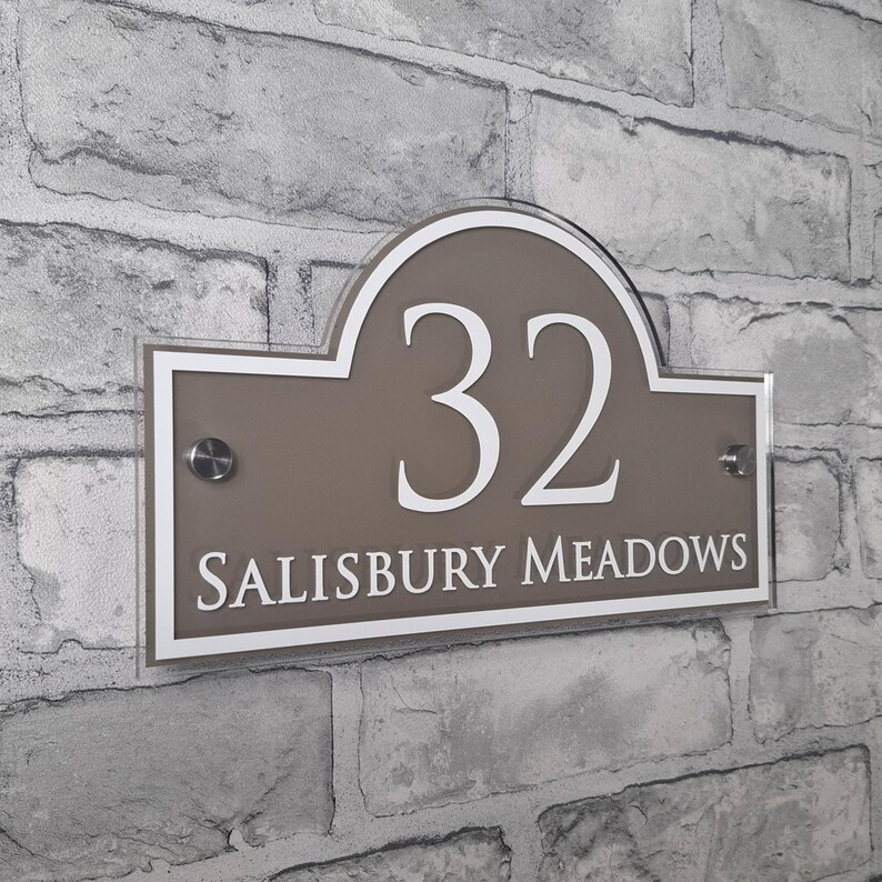 May include: A rectangular house number sign with the number "32" in white, above the words "SALISBURY MEADOWS". The sign is taupe with a white border and is mounted on a brick wall.