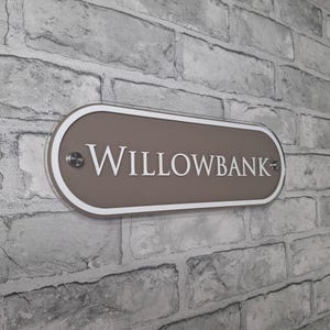 May include: A rectangular, oval-shaped sign with the word "WILLOWBANK" in white lettering. The sign has a taupe center, a white border, and is mounted on a brick wall. The sign is secured with two silver-colored screws.