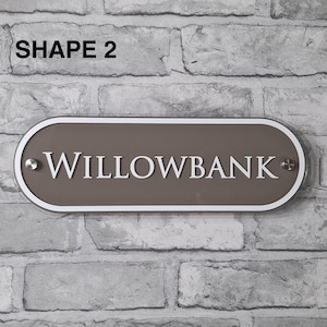 May include: A rectangular, oval-shaped sign with the text "WILLOWBANK" in white lettering. The sign has a brown center, a white border, and is mounted on a brick wall. The text "SHAPE 2" is in the top left corner.