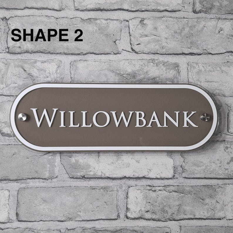 May include: A rectangular, oval-shaped sign with the text "WILLOWBANK" in white lettering. The sign has a brown center, a white border, and is mounted on a brick wall. The text "SHAPE 2" is in the top left corner.