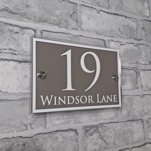 May include: Rectangular house number sign with the number "19" in white, and the words "Windsor Lane" in white, on a taupe background. The sign is framed in white and mounted on a brick wall.