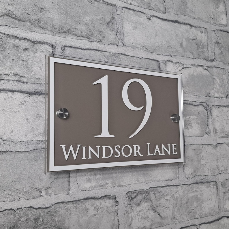 May include: Rectangular house number sign with the number "19" in white, and the words "Windsor Lane" in white, on a taupe background. The sign is framed in white and mounted on a brick wall.