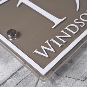 May include: A rectangular house sign with the number 1 and the name "Windsor" in white lettering. The sign has a taupe background with a white border and a silver-colored mounting fixture.