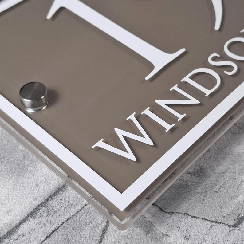 May include: A rectangular house sign with the number 1 and the name "Windsor" in white lettering. The sign has a taupe background with a white border and a silver-colored mounting fixture.