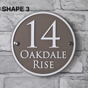 May include: A round, brown house number sign with the number "14" in white, along with the text "OAKDALE RISE". The sign has a white border and is mounted on a brick wall. The text "SHAPE 3" is at the top.