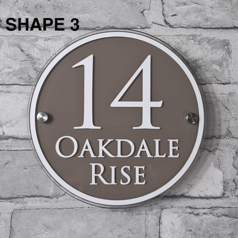 May include: A round, brown house number sign with the number "14" in white, along with the text "OAKDALE RISE". The sign has a white border and is mounted on a brick wall. The text "SHAPE 3" is at the top.