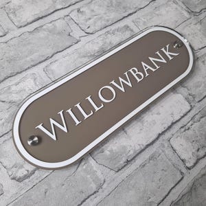 May include: A rectangular sign with rounded edges, featuring the word "WILLOWBANK" in white capital letters. The sign has a taupe background with a white border and is mounted with silver-colored hardware. The sign is displayed against a gray brick wall.