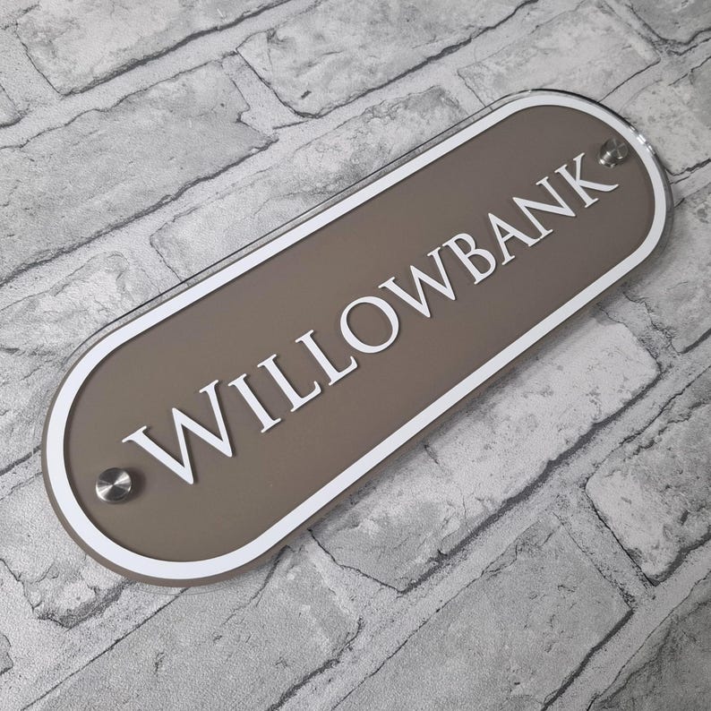 May include: A rectangular sign with rounded edges, featuring the word "WILLOWBANK" in white capital letters. The sign has a taupe background with a white border and is mounted with silver-colored hardware. The sign is displayed against a gray brick wall.